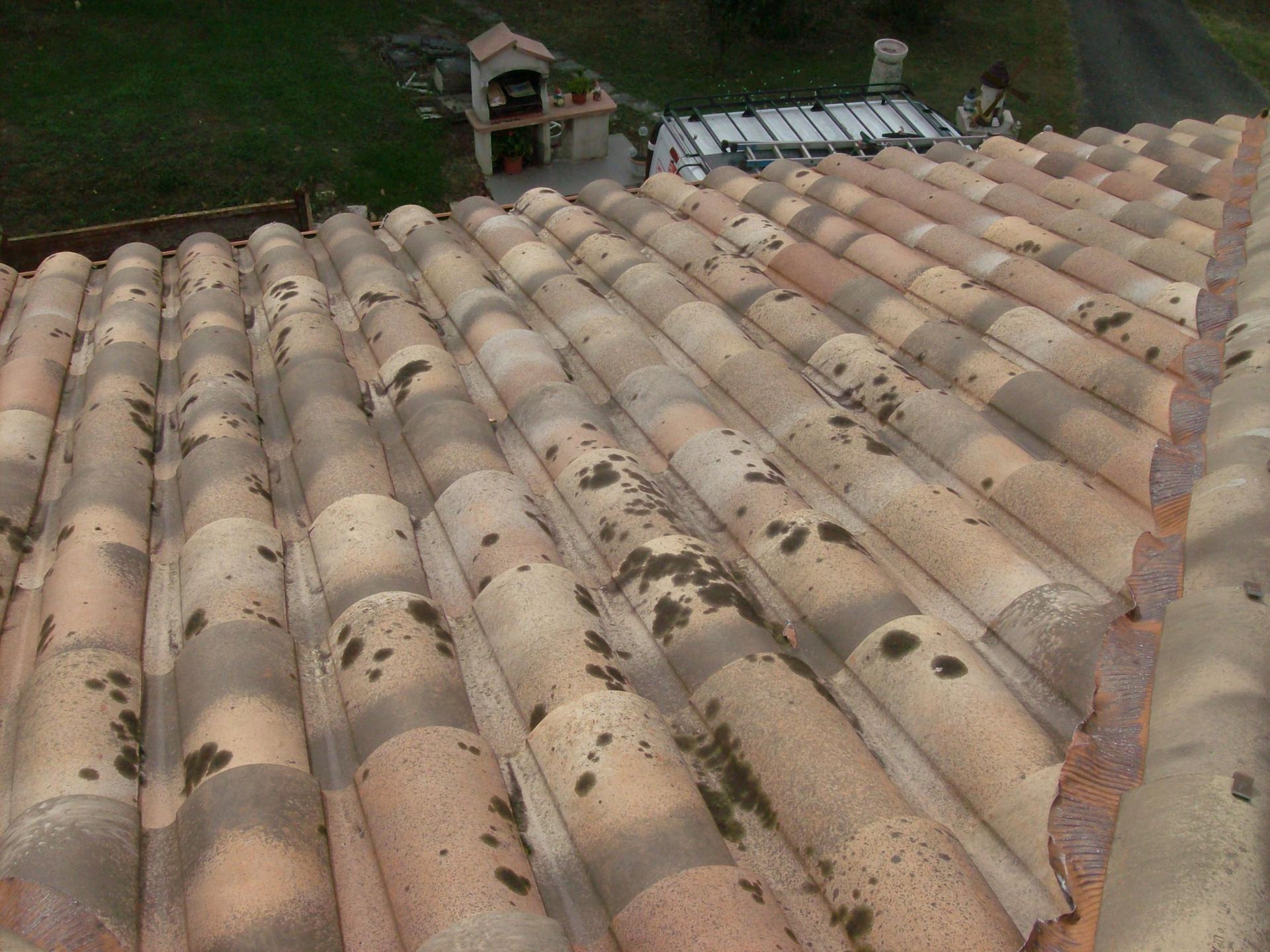 Gros plan sur un toit en tuiles de terre cuite parsemé de mousse, donnant sur une allée et un jardin.