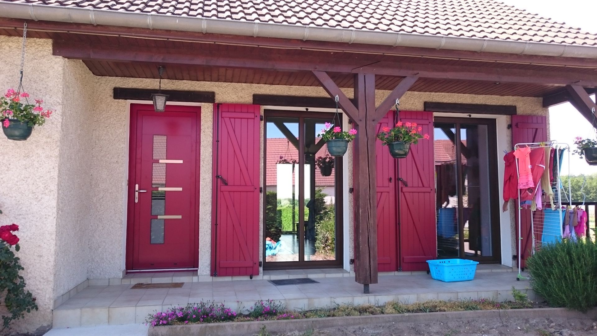 Porte rouge et volets sur une maison, sous un porche couvert, avec des paniers de fleurs et une corde à linge visibles.