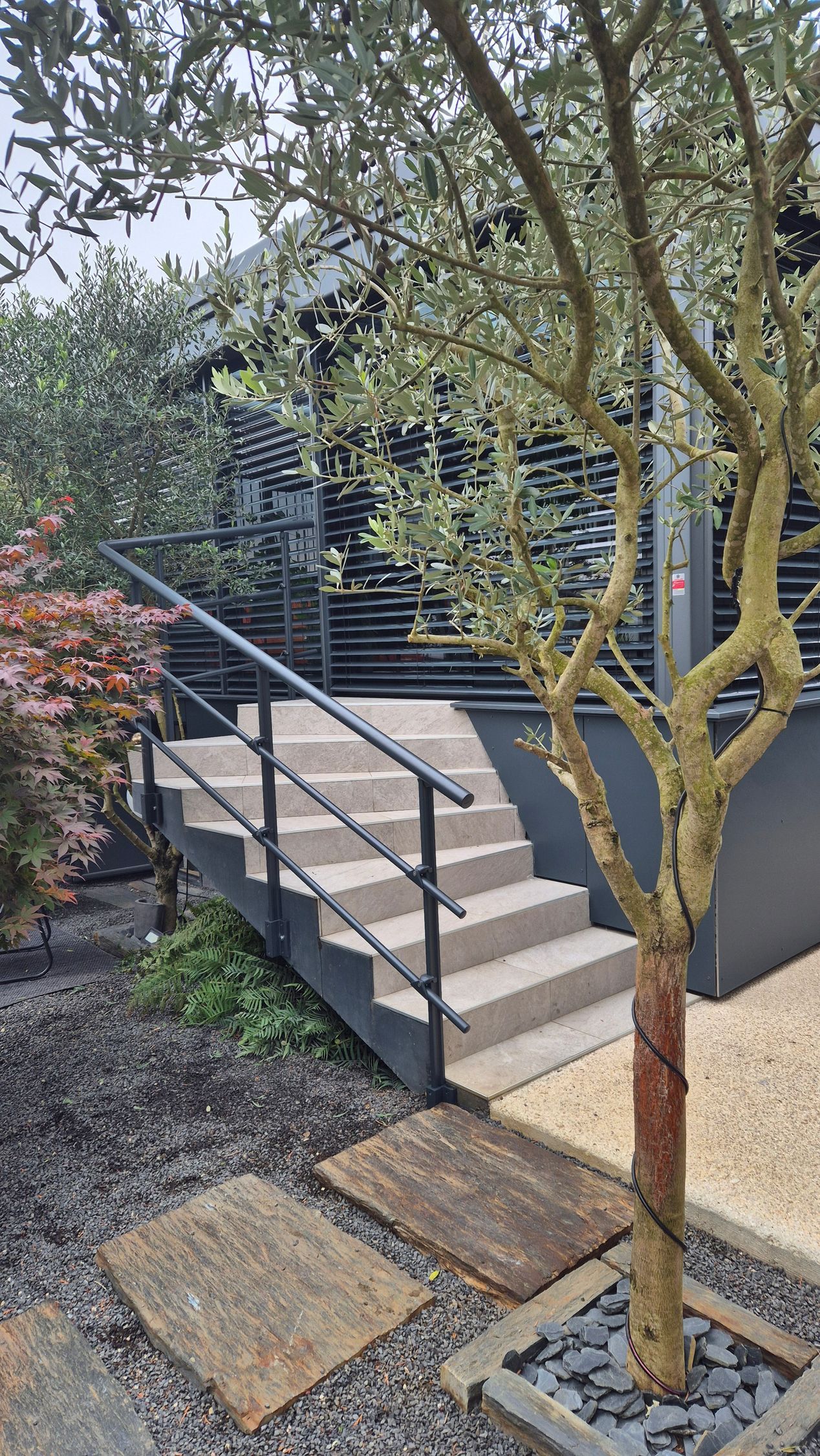 Escalier extérieur avec rampe noire menant à un bâtiment sombre ; un arbre au premier plan.