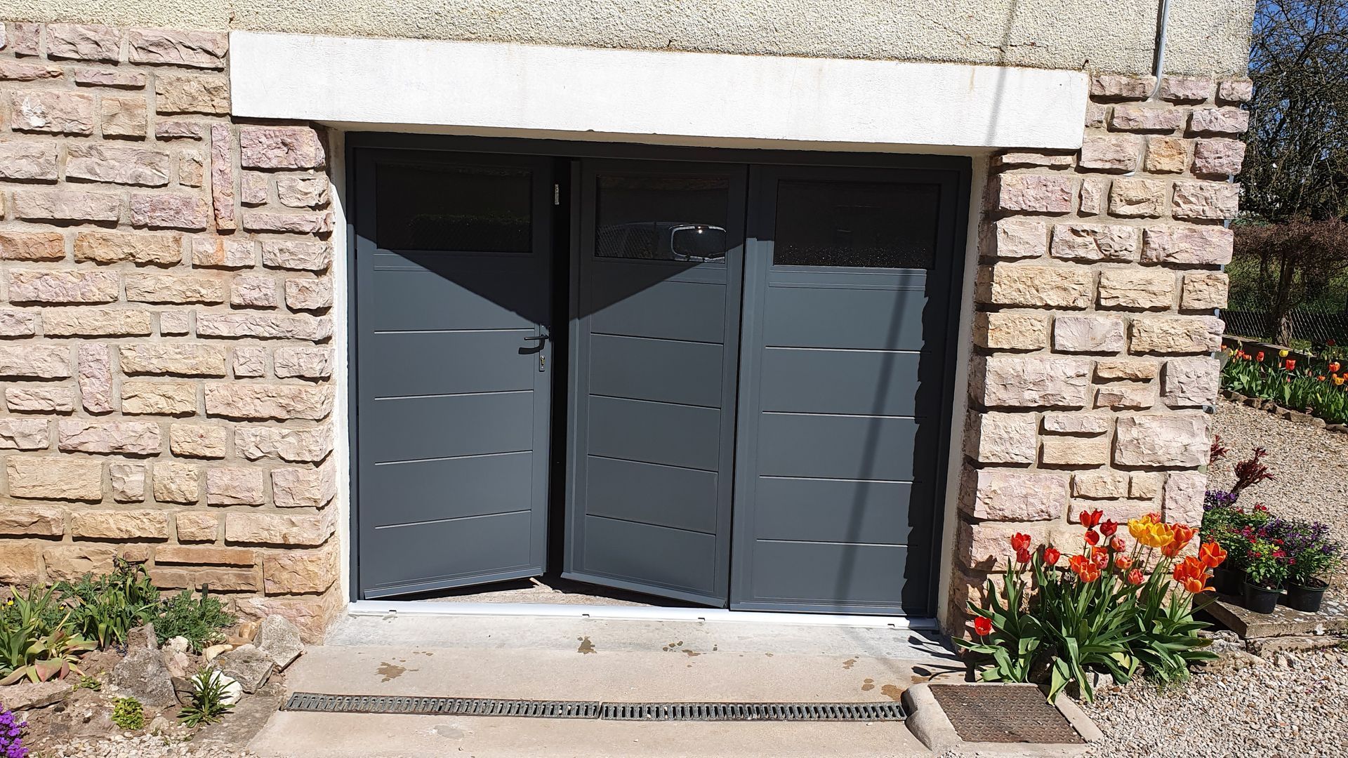 Portes de garage grises, entrouvertes, encastrées dans un mur de pierre. Fleurs et plantes encadrent l'entrée.