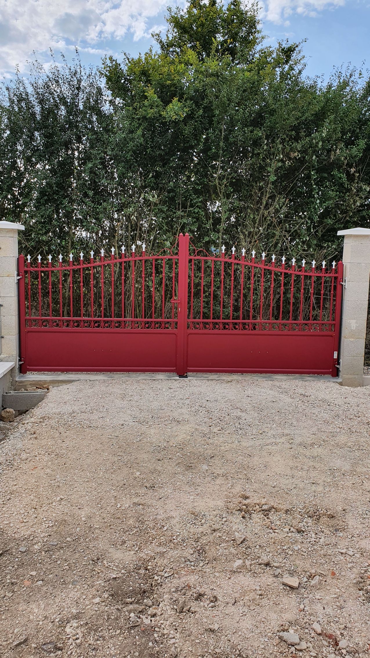 Un portail en métal rouge entre deux piliers de pierre, situé dans une allée de gravier avec des arbres verts en arrière-plan.