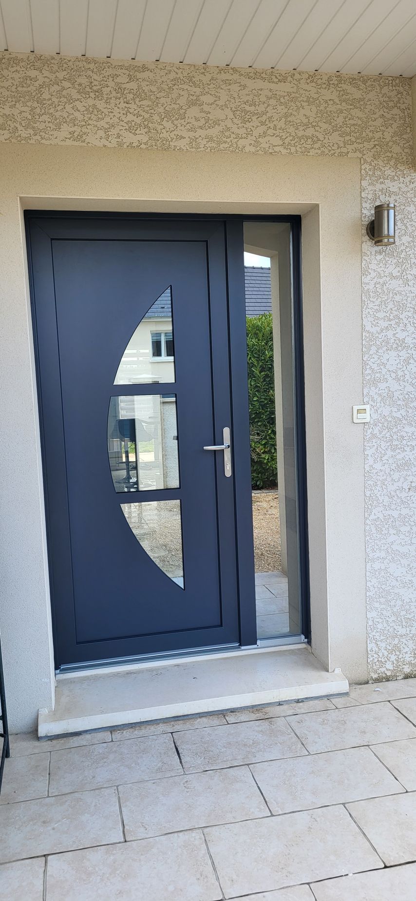 Une porte d'entrée bleu foncé avec une fenêtre ovale. Un panneau latéral transparent. Des murs et un sol en pierre beige.