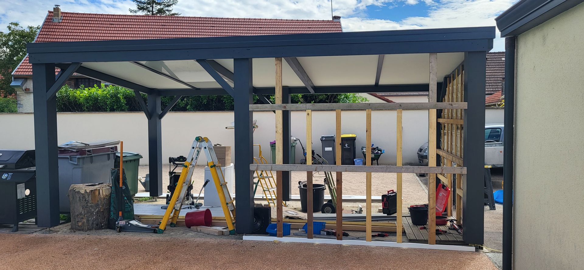 Dans un jardin, on aperçoit une pergola grise partiellement construite, avec une structure en bois, des outils et des matériaux de construction.