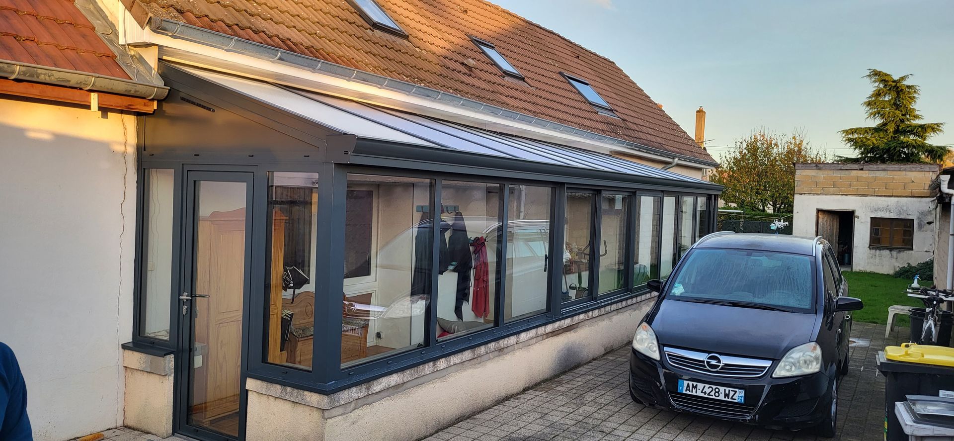 Une maison avec une véranda vitrée jouxte une voiture noire garée sur une allée pavée.