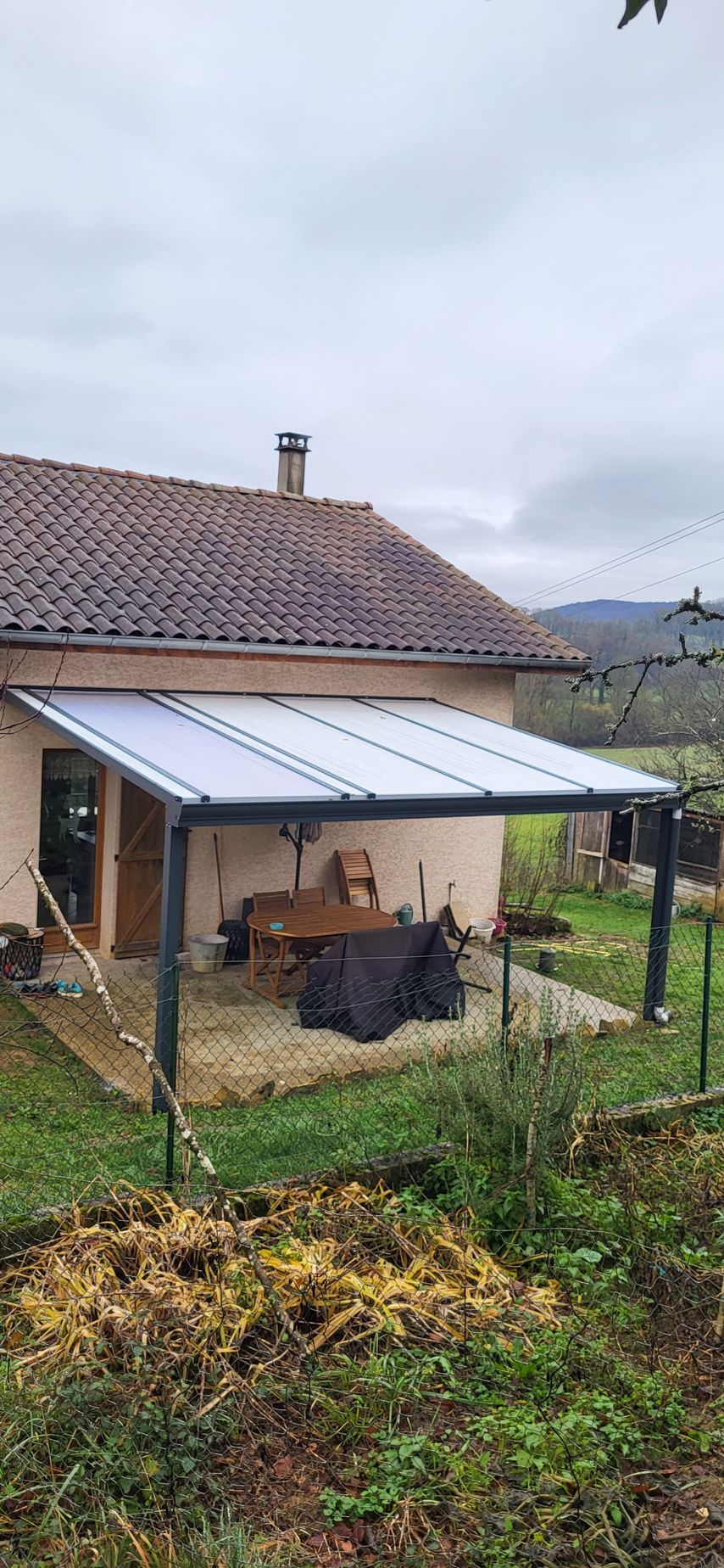Une maison avec un patio couvert d'un toit blanc, entourée d'une cour avec de l'herbe verte et des feuilles brunes.