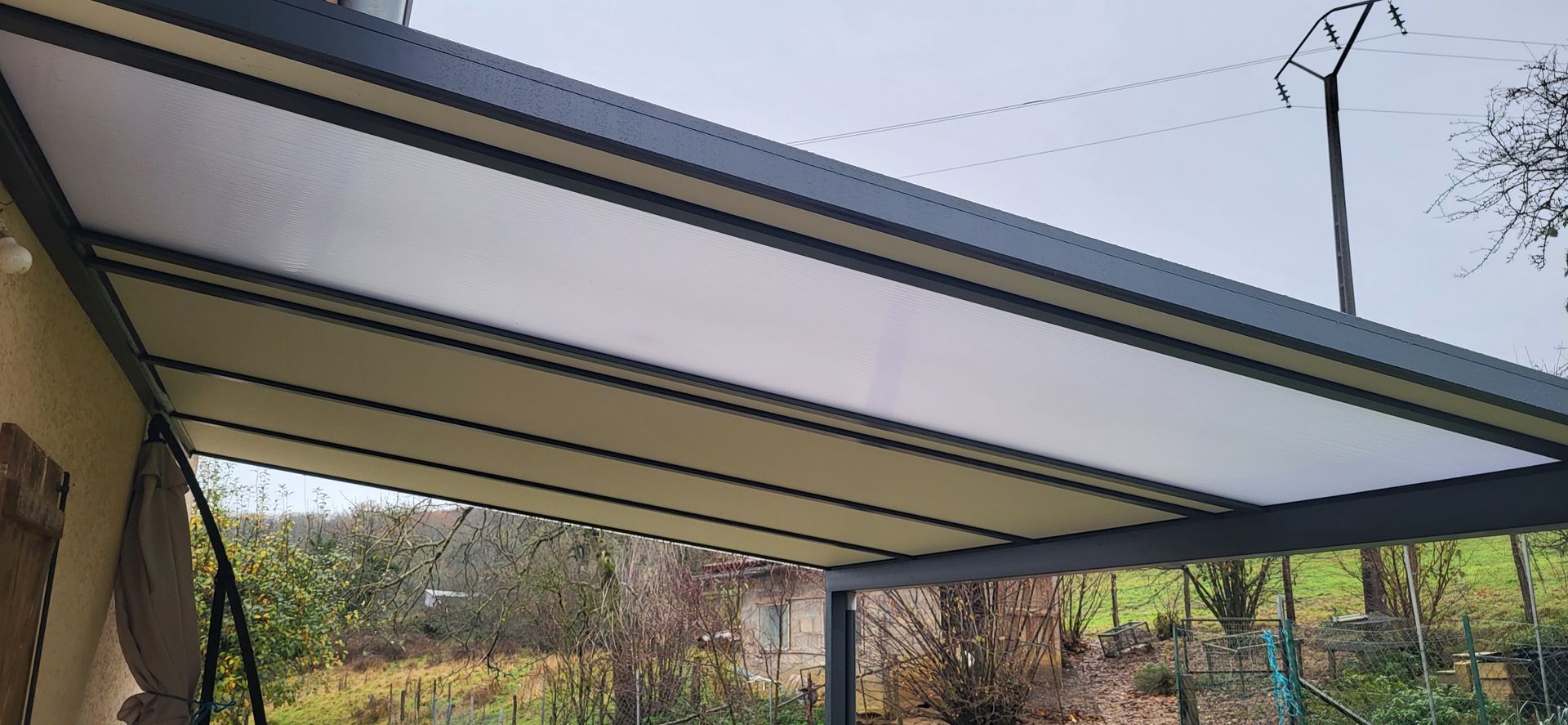 Un toit de patio moderne avec des panneaux blancs rétractables, entouré de verdure et d'un ciel couvert.