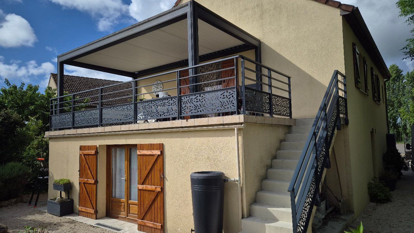 Maison beige à deux étages avec balcon et pergola en métal, volets en bois, escalier extérieur.