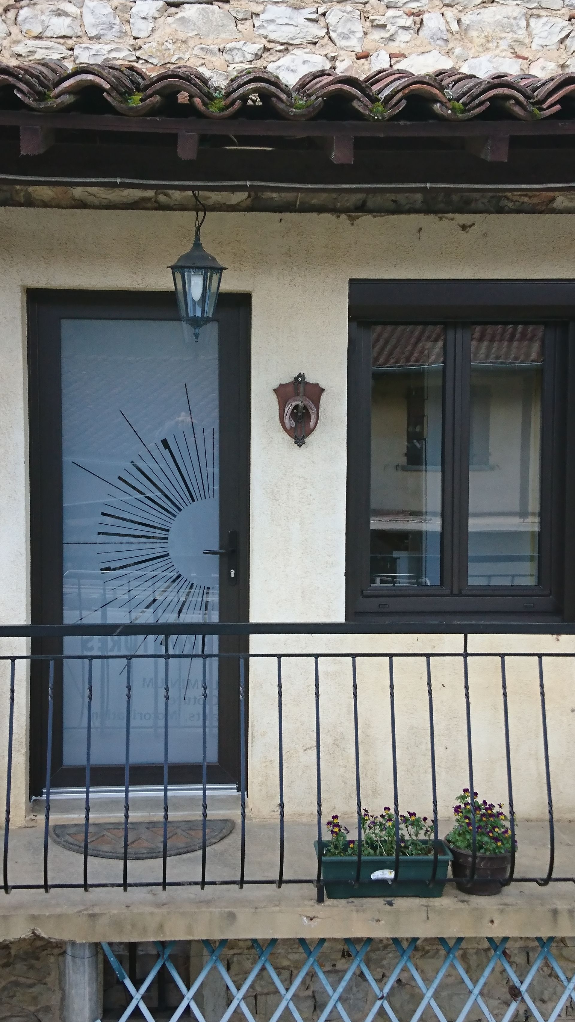 Vue extérieure d'un bâtiment avec une porte et une fenêtre, une applique murale ornée, un petit balcon fleuri et un motif de soleil rayonnant sur la porte.