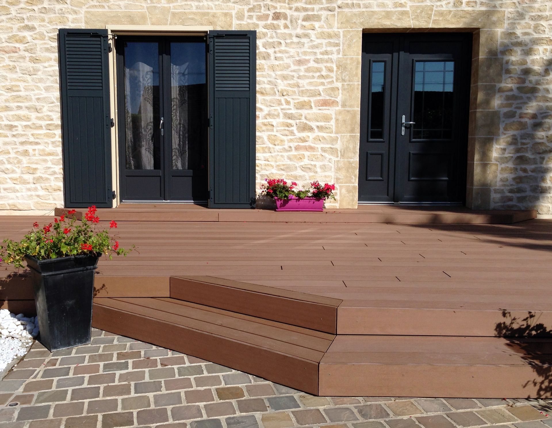 Terrasse et marches en composite marron menant à des portes et volets noirs, façade en pierre.