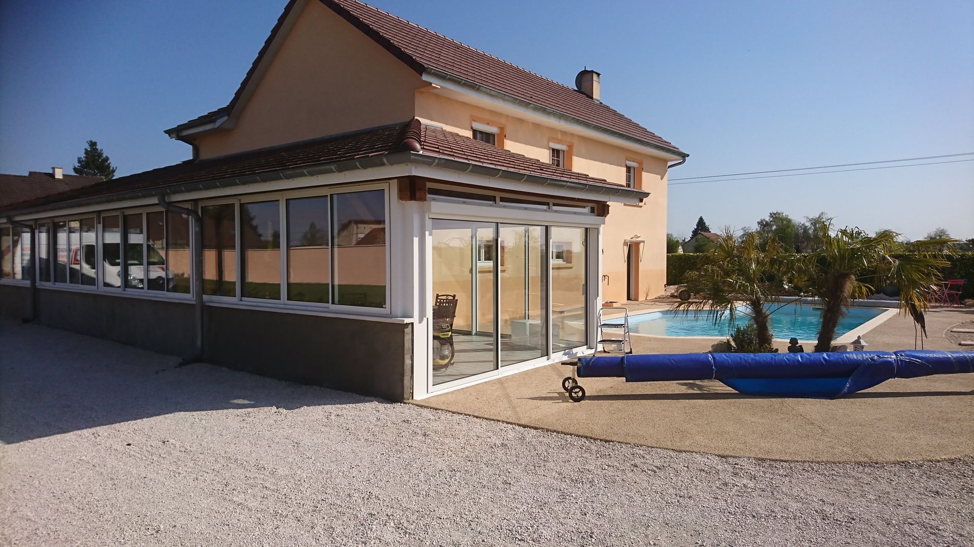 Maison beige avec un patio vitré donnant sur une piscine. Allée de gravier au premier plan.