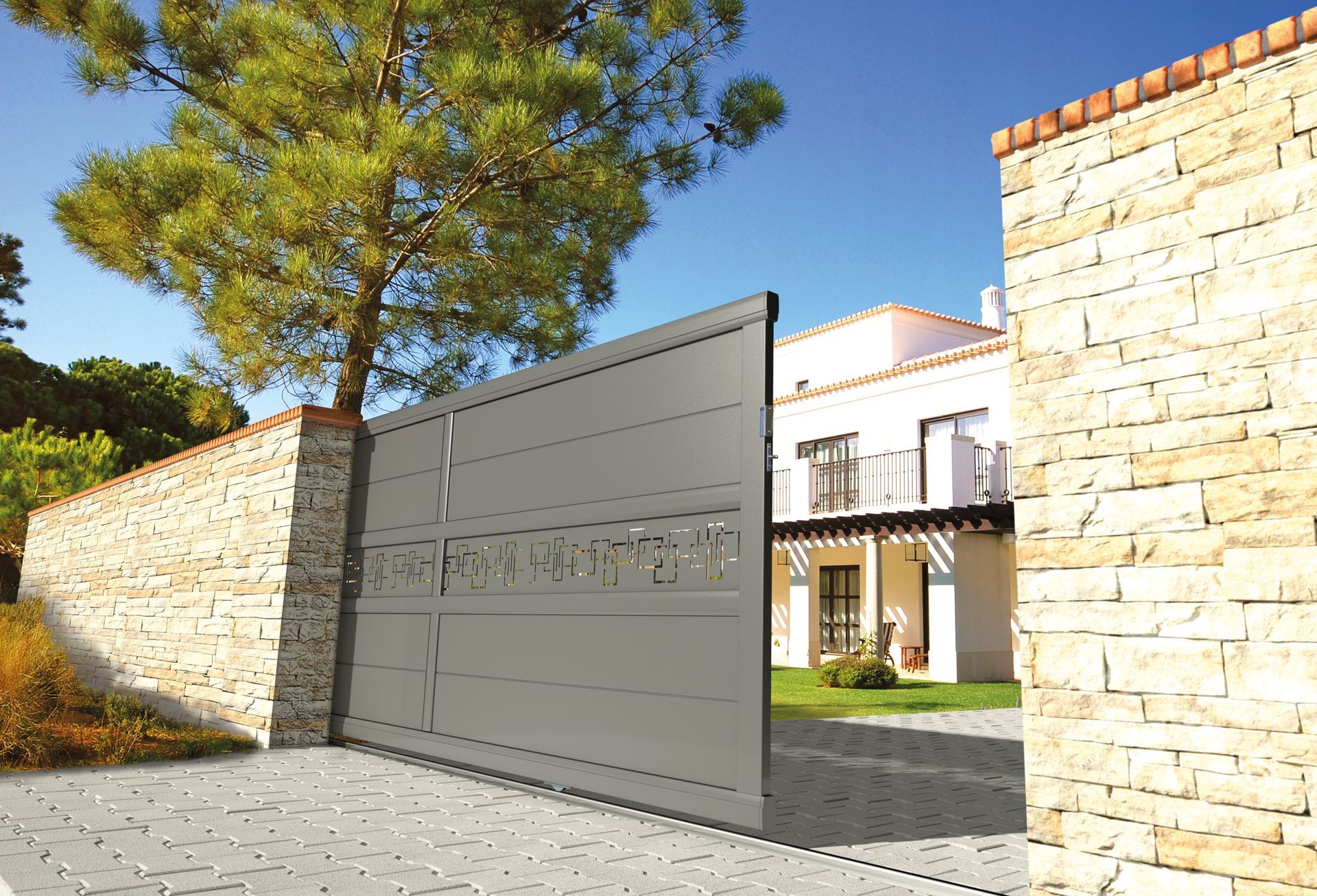 Portail gris orné de découpes décoratives, encastré dans un mur de pierre, donnant sur une maison avec balcon.