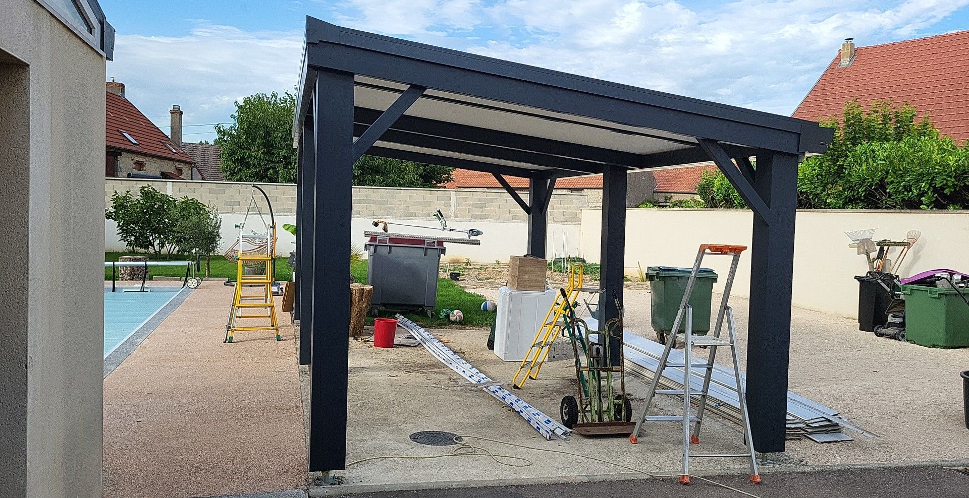 Pergola en construction dans une cour gravillonnée, avec des outils et une piscine visibles.