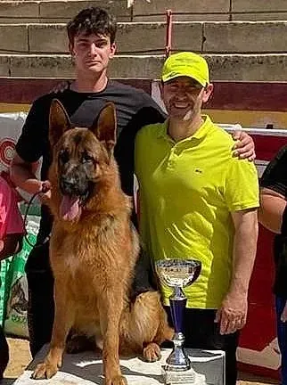 Dos hombres y un pastor alemán posan para una foto con un trofeo.