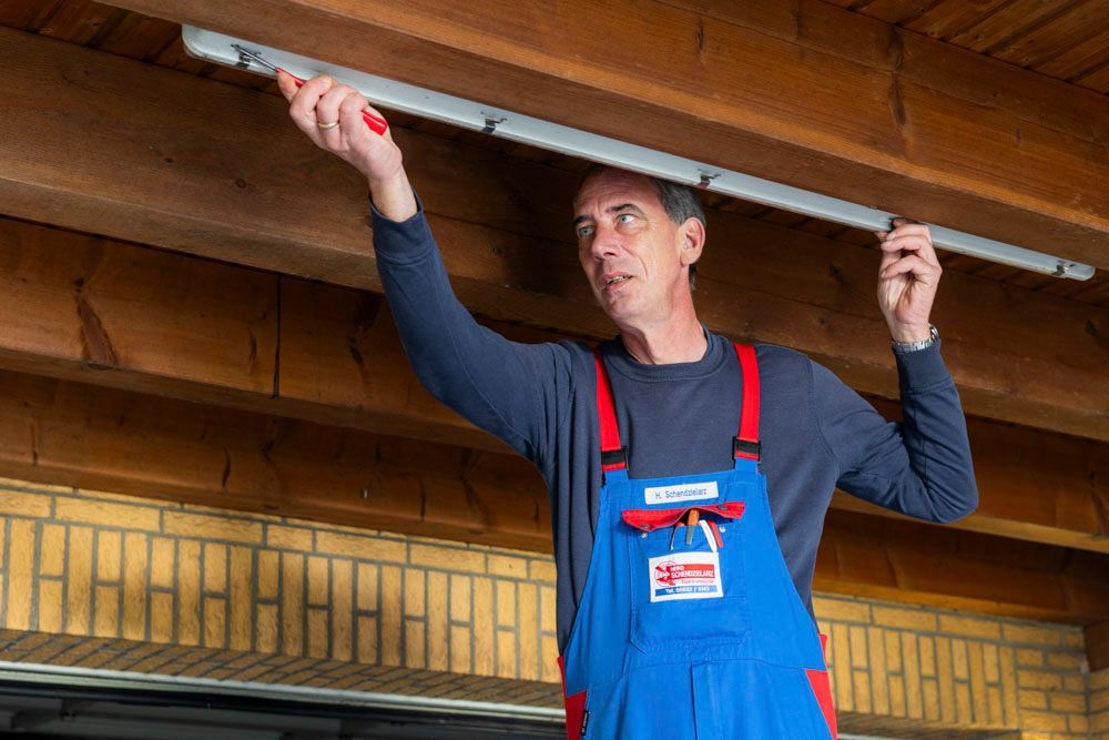 Ein Mann in blauem Overall montiert eine Lampe an einem Holzbalken und hält dabei ein Werkzeug in der Hand.