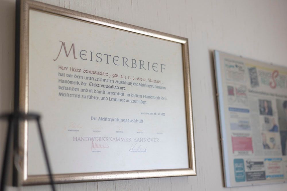 Ein gerahmtes Dokument mit dem Wort „Meisterbrief“ an einer weißen Wand, im Hintergrund ein weiteres gerahmtes Objekt.