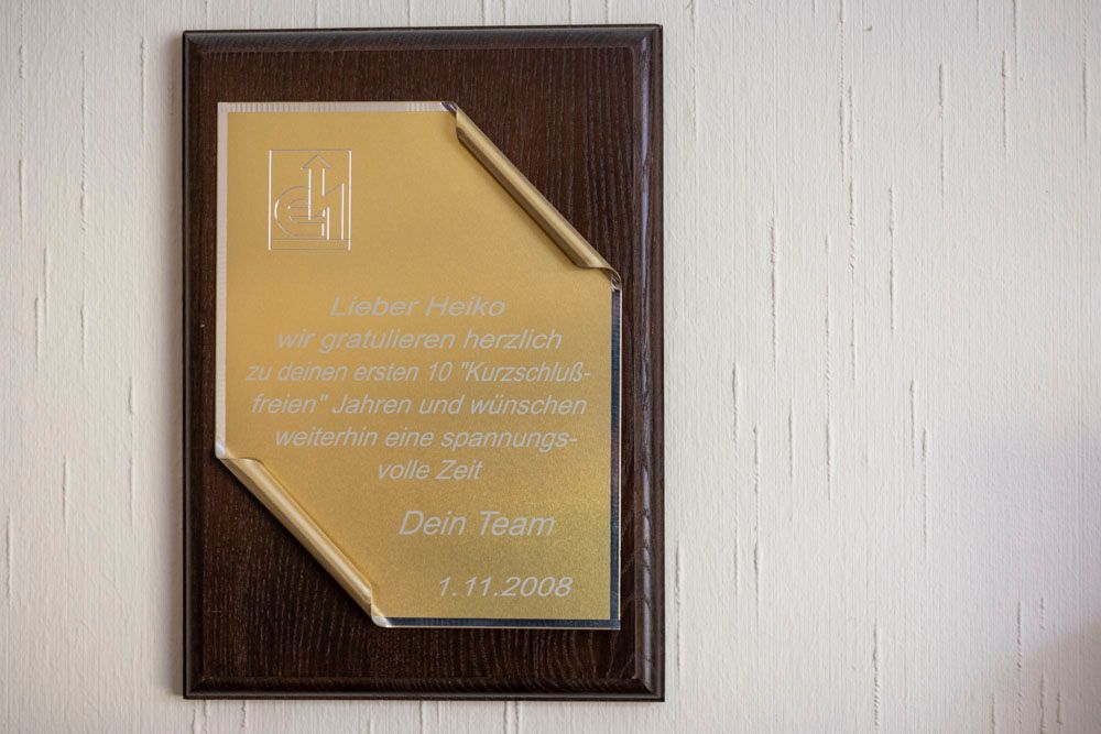 Plakette mit Goldplatte und Text auf Holzgrund, an einer weißen Wand angebracht.