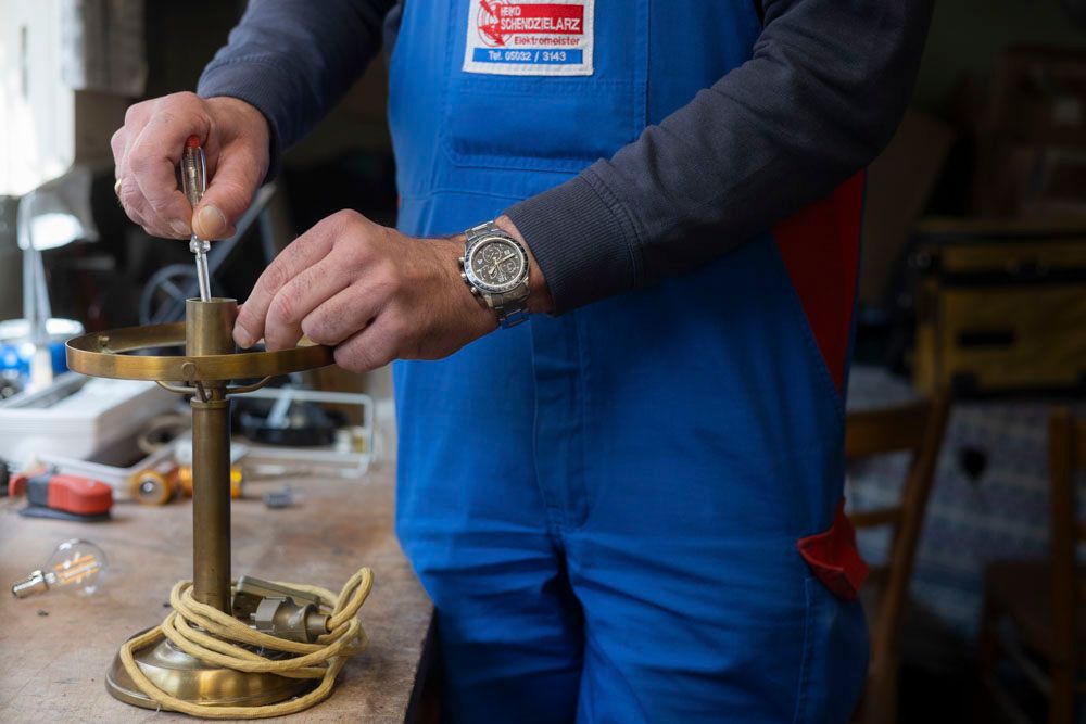 Eine Person in blauem Overall repariert mit einem Schraubenzieher eine Lampe in einem Innenraum.