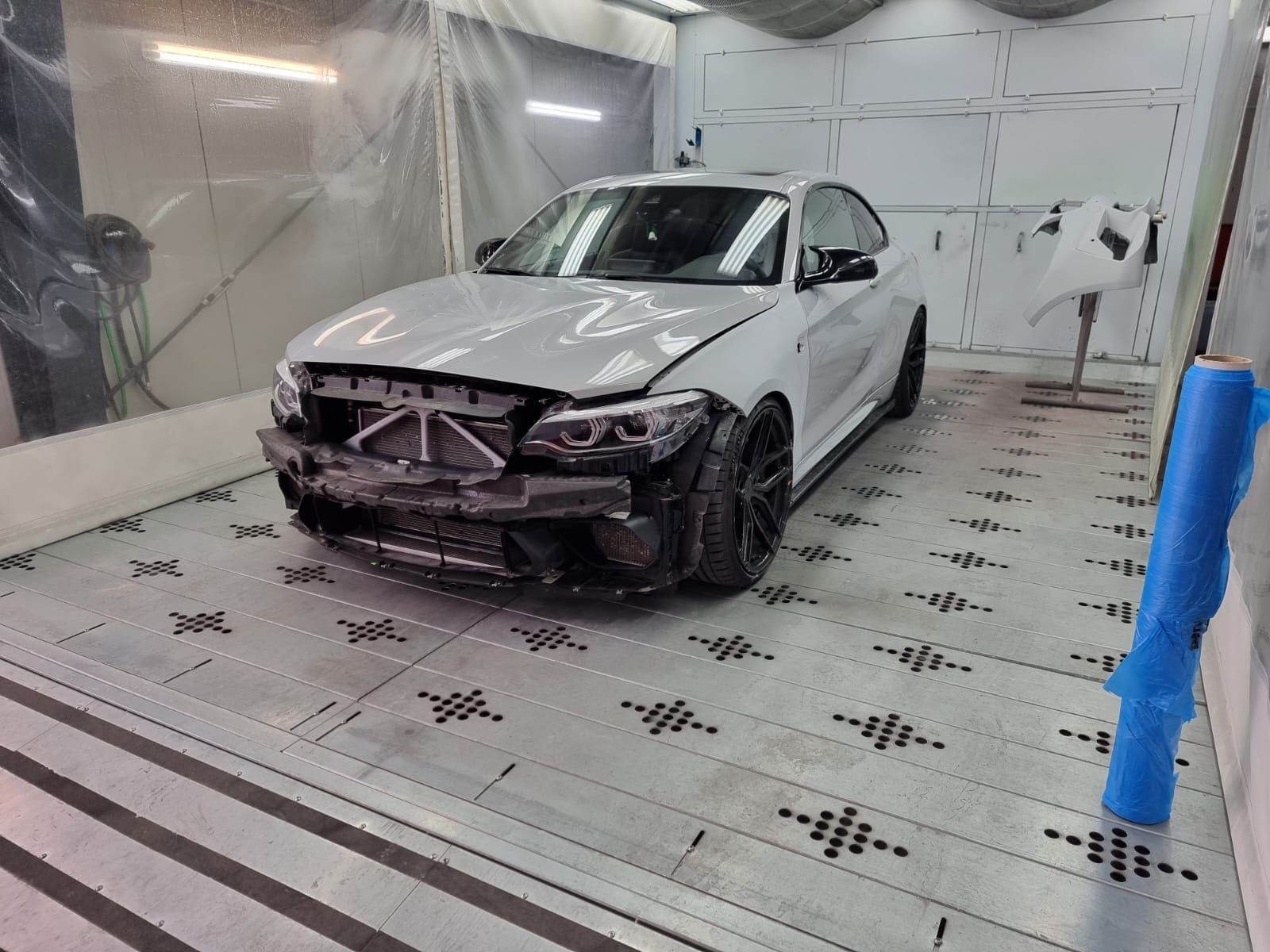 Berline blanche endommagée dans une cabine de carrosserie, pare-chocs et calandre avant démontés.