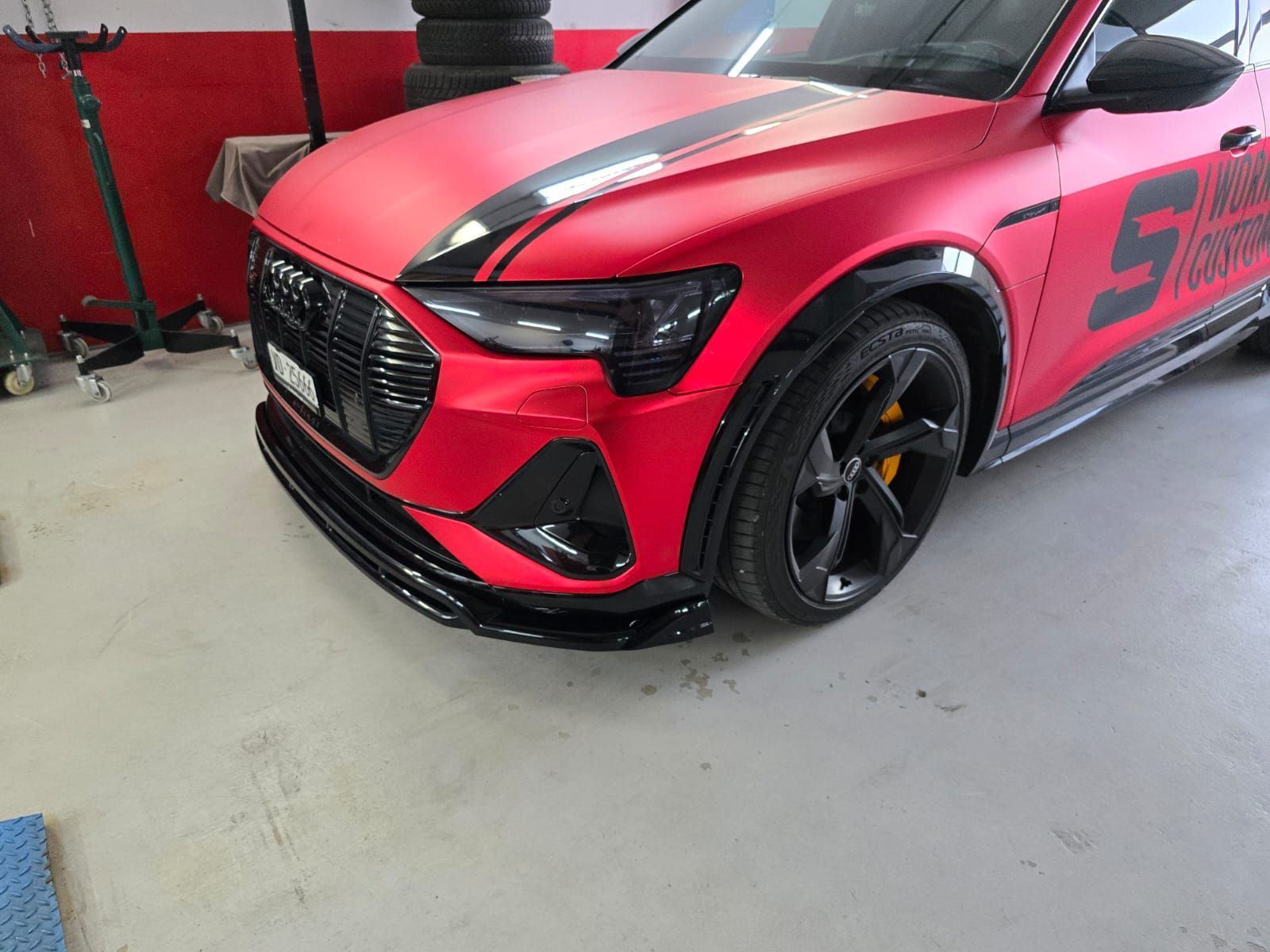 Berline sport rouge dans un garage, vue de face gauche, avec bandes de course noires et grandes roues noires.