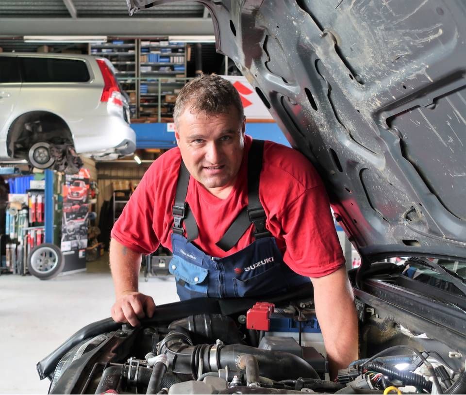Ein Mechaniker in rotem Hemd und Overall arbeitet in einer Garage unter der Motorhaube eines Autos.