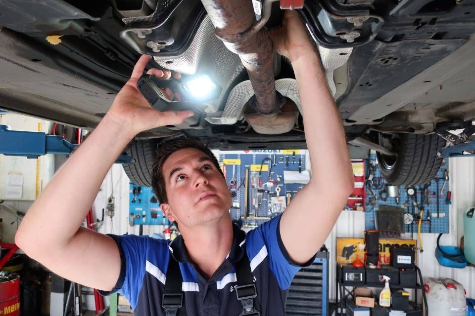 Ein Mechaniker untersucht in einer Autowerkstatt mit einer Taschenlampe die Unterseite eines Autos.