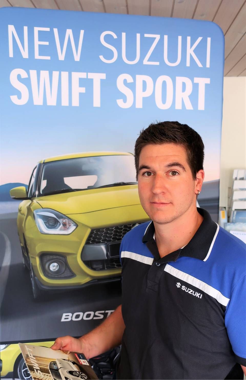 Ein Mann steht neben einem Poster eines gelben neuen Suzuki Swift Sport an einem Messestand.