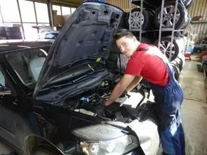 Ein Mechaniker im roten Hemd arbeitet in einer Autowerkstatt unter der Motorhaube eines Wagens.