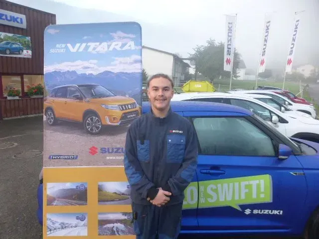 Mann steht neben einem blauen Suzuki Swift und einem Vitara-Ausstellungsstück in einem Autohaus.