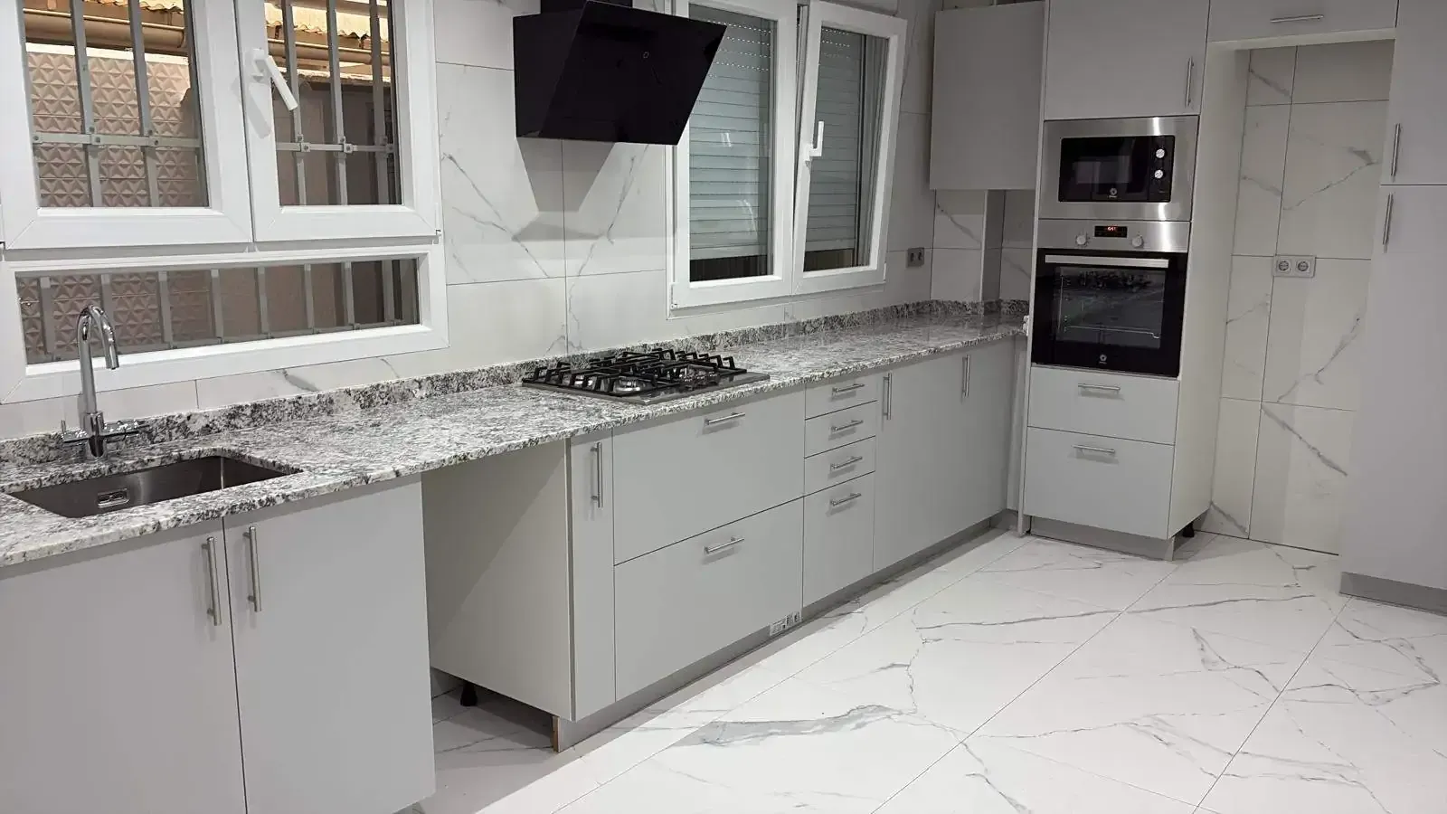 Cocina moderna blanca con encimeras de granito, electrodomésticos integrados y grandes ventanales.