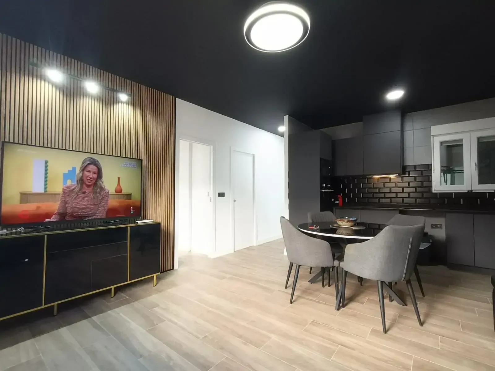 Cocina y comedor modernos de planta abierta con techo negro, pared decorativa de madera y televisor montado en la pared.