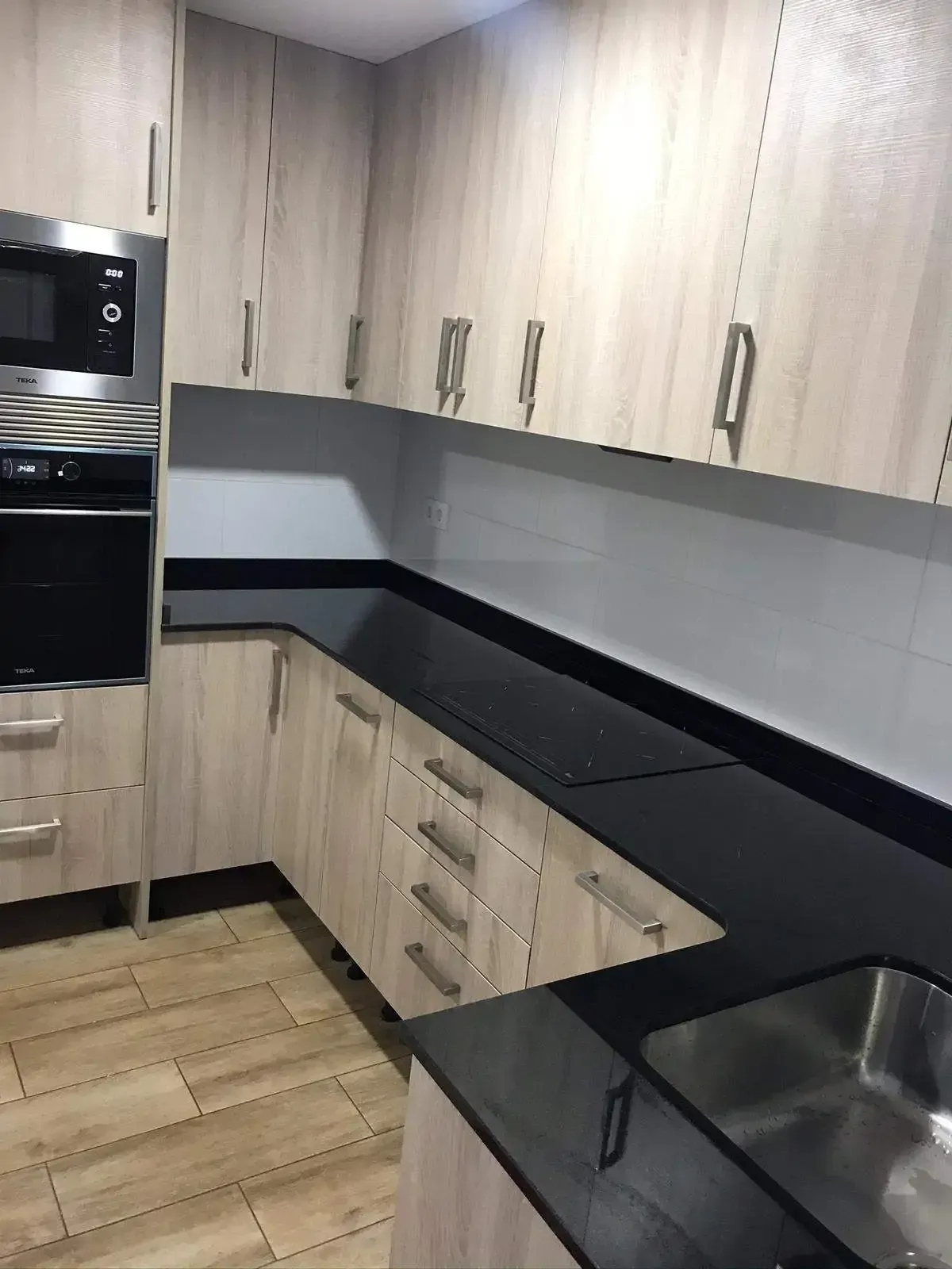 Cocina moderna con armarios de madera clara, encimeras negras y fregadero de acero inoxidable.