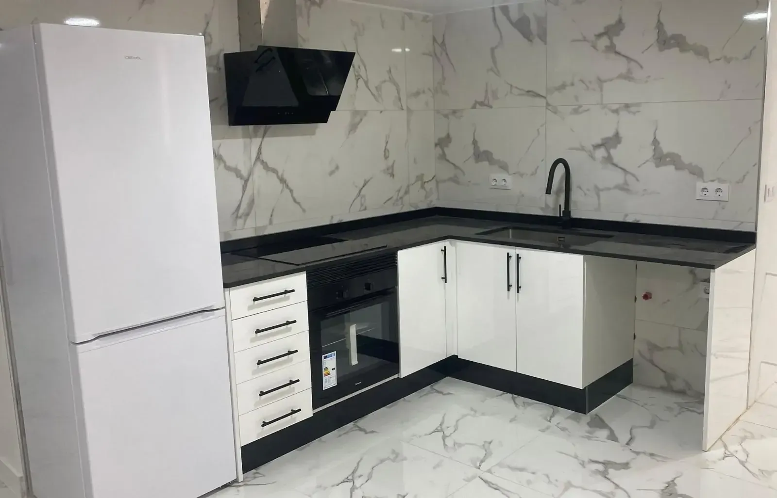 Cocina moderna en forma de L con armarios blancos, encimeras negras, paredes con estampado de mármol y un frigorífico blanco.