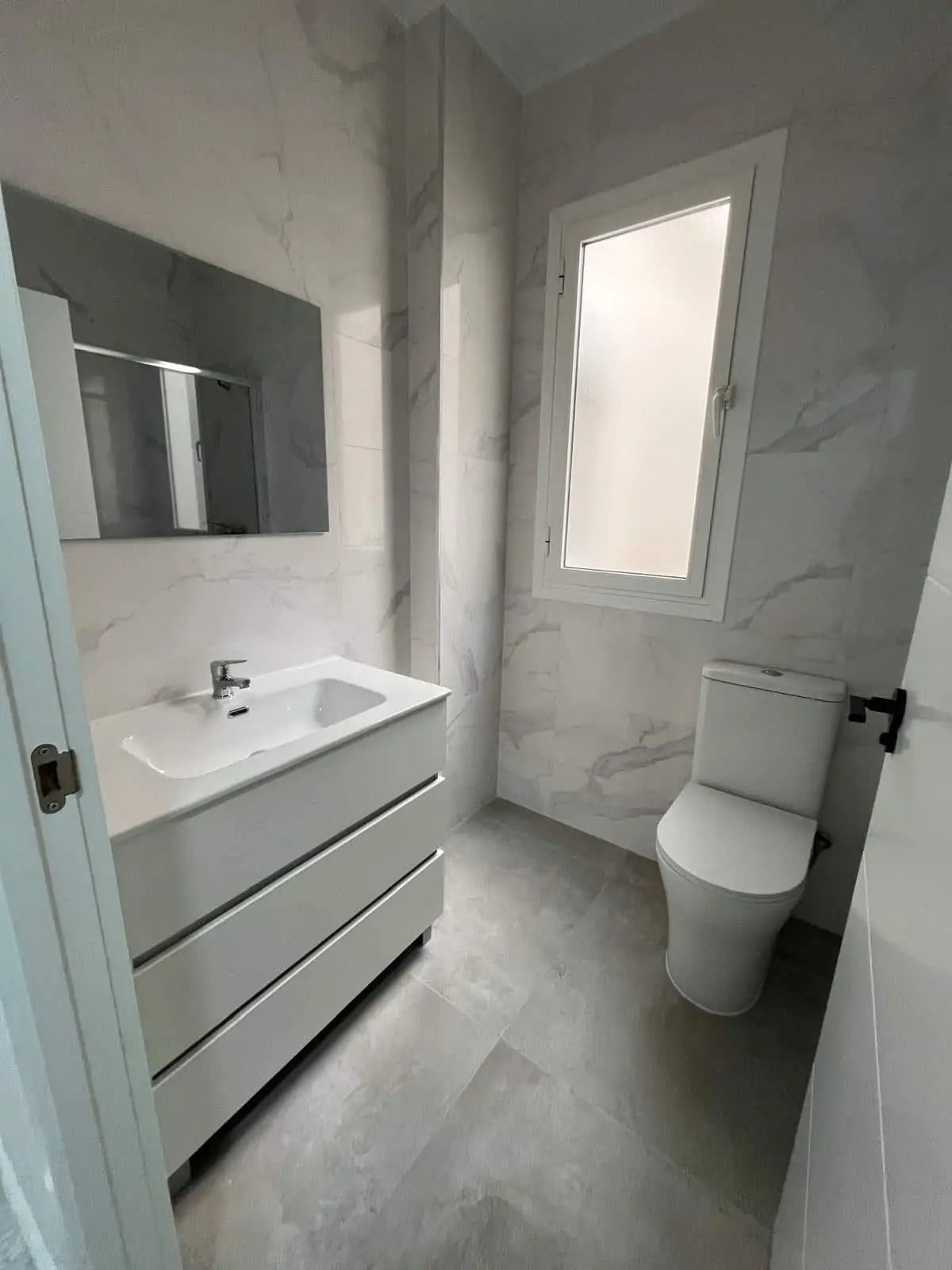 Baño moderno blanco con tocador, espejo, inodoro y ventana esmerilada en una habitación luminosa con azulejos.
