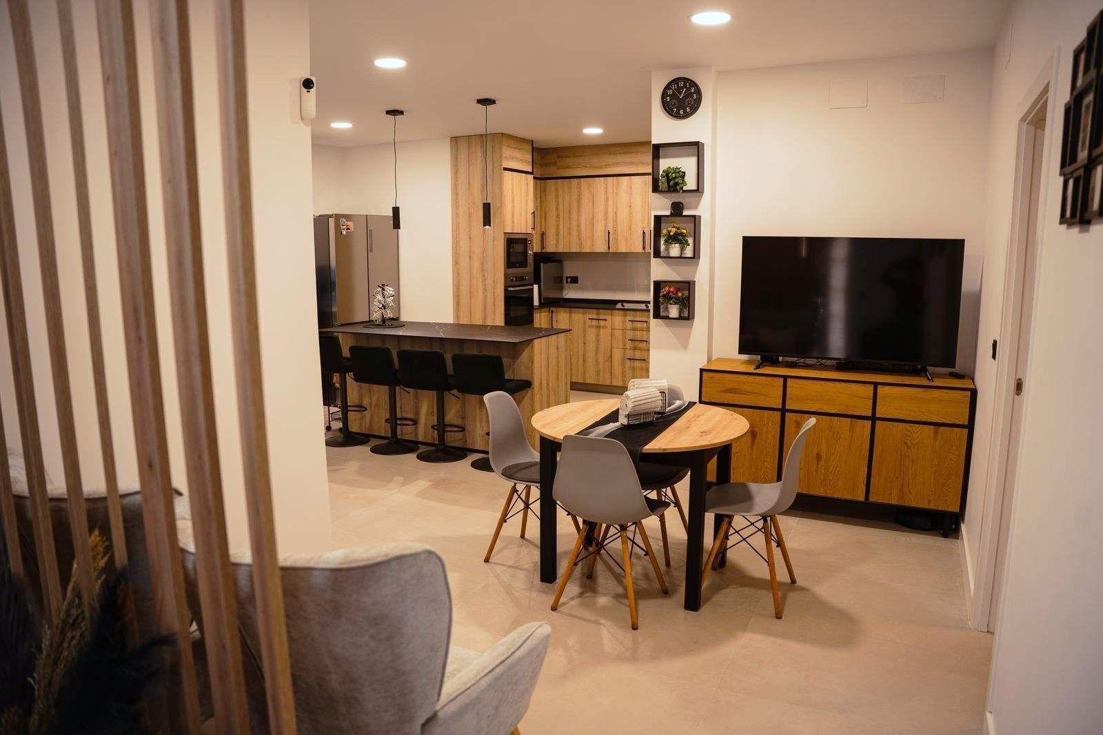 Salón-cocina moderno de planta abierta con mesa de comedor, sofá gris y televisor montado en la pared.
