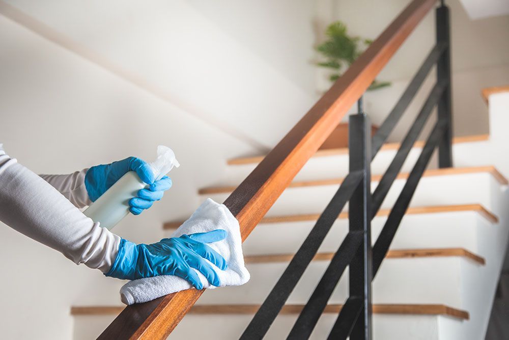 Eine Person mit blauen Handschuhen desinfiziert ein hölzernes Treppengeländer mit Spray und Tuch.