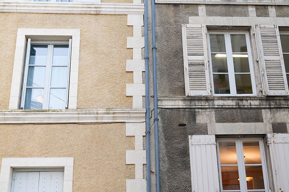 Zwei nebeneinanderliegende Gebäude; eines aus hellbraunem Stuck, das andere aus grauem Stein. Fenster sind sichtbar, einige mit Fensterläden.
