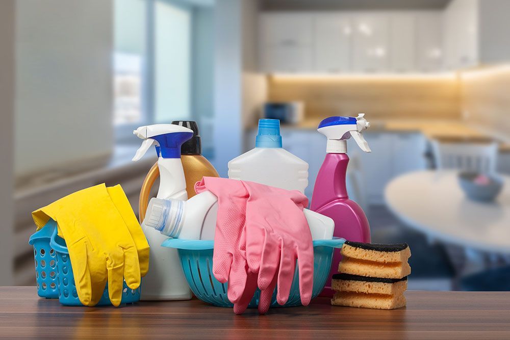 Reinigungsmittel auf einem Tisch: Sprühflaschen, Handschuhe, Schwämme und ein Korb, in einer Küchenumgebung.