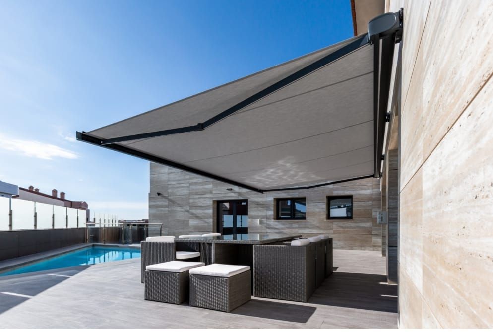 Patio exterior moderno con toldo retráctil gris, conjunto de muebles de mimbre y piscina adosada a un muro de piedra.