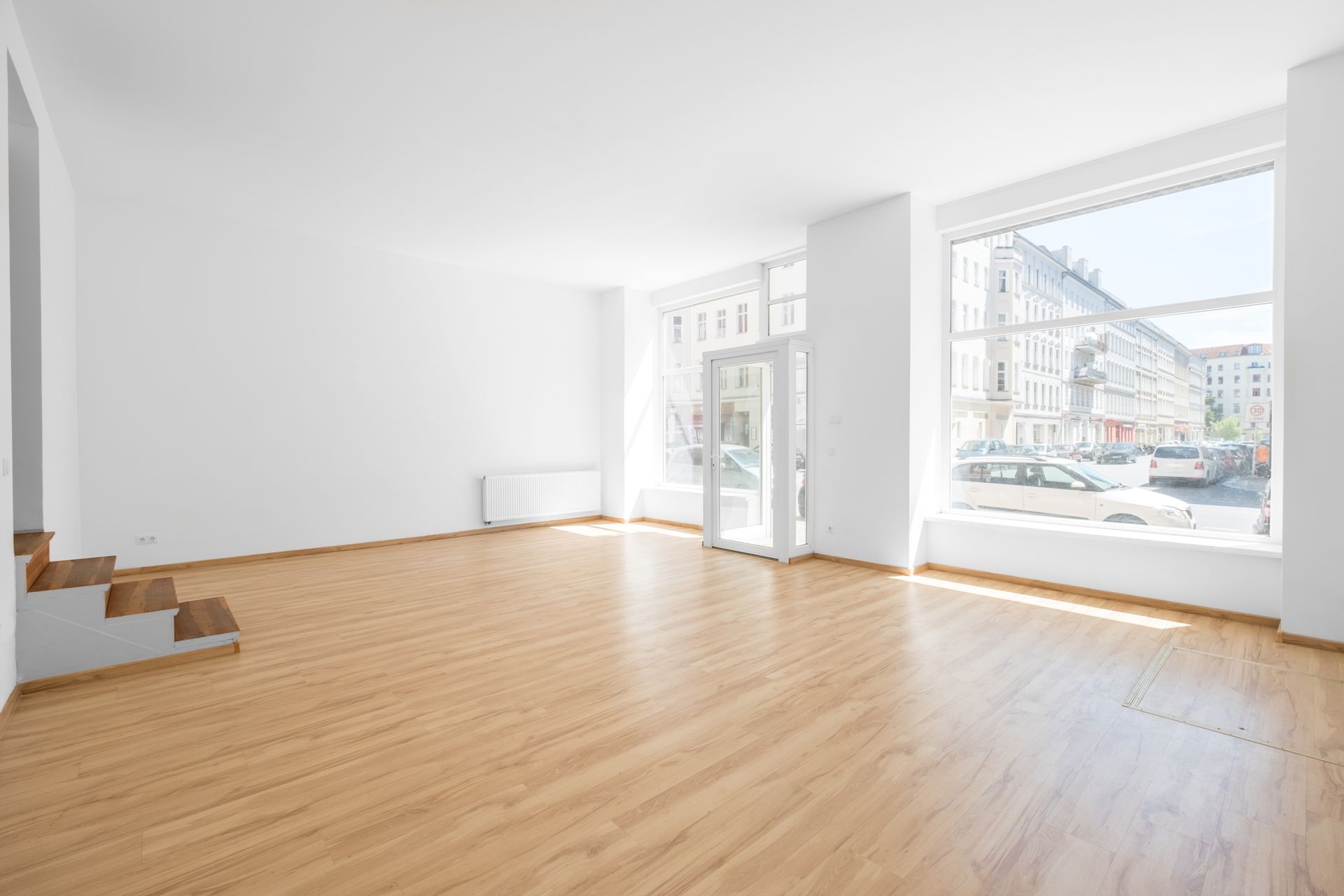 Pièce lumineuse et vide, avec un parquet, des murs blancs et de grandes fenêtres donnant sur une rue de la ville