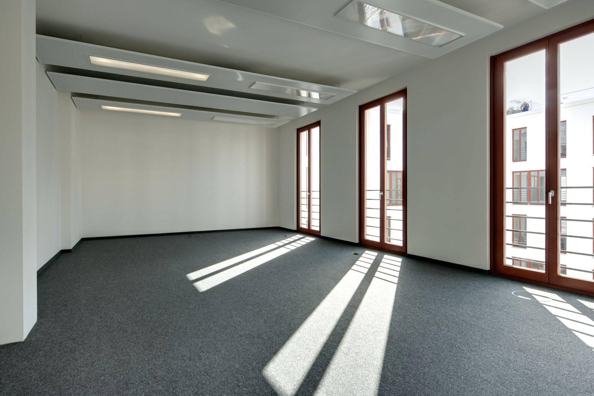 Bureau vide baigné de soleil, avec des murs blancs, une moquette grise et de grandes fenêtres projetant de longues ombres