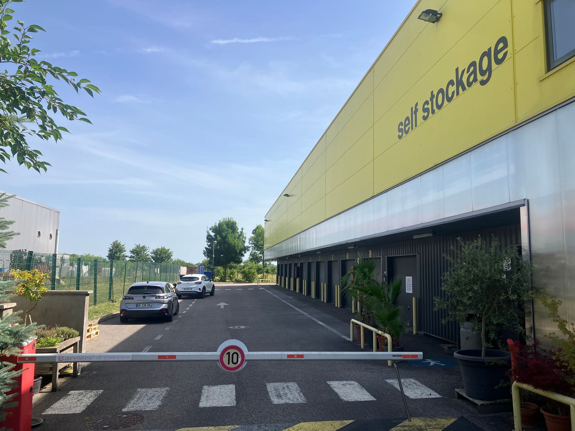 Entrée d’un centre d'entreposage en libre-service avec des voitures en stationnement, un bâtiment jaune et une barrière.