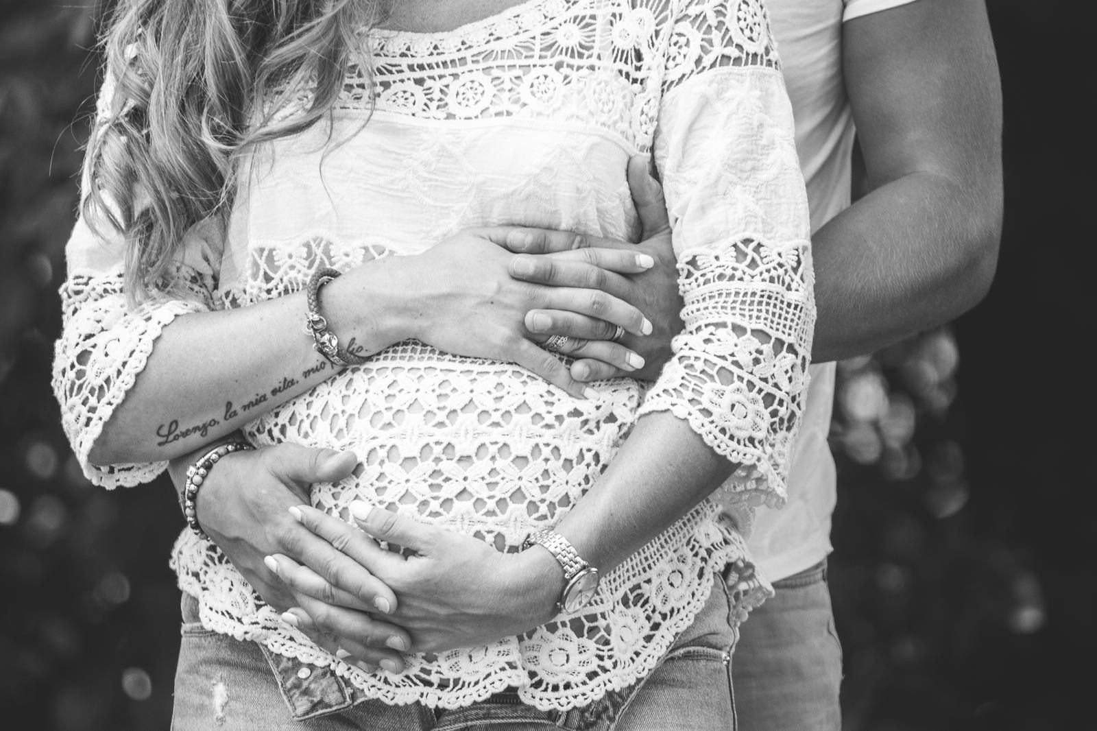 Une femme enceinte, vêtue d'un haut en dentelle, tient son ventre à deux mains tandis qu'une autre personne se tient derrière elle, les bras autour d'elle.