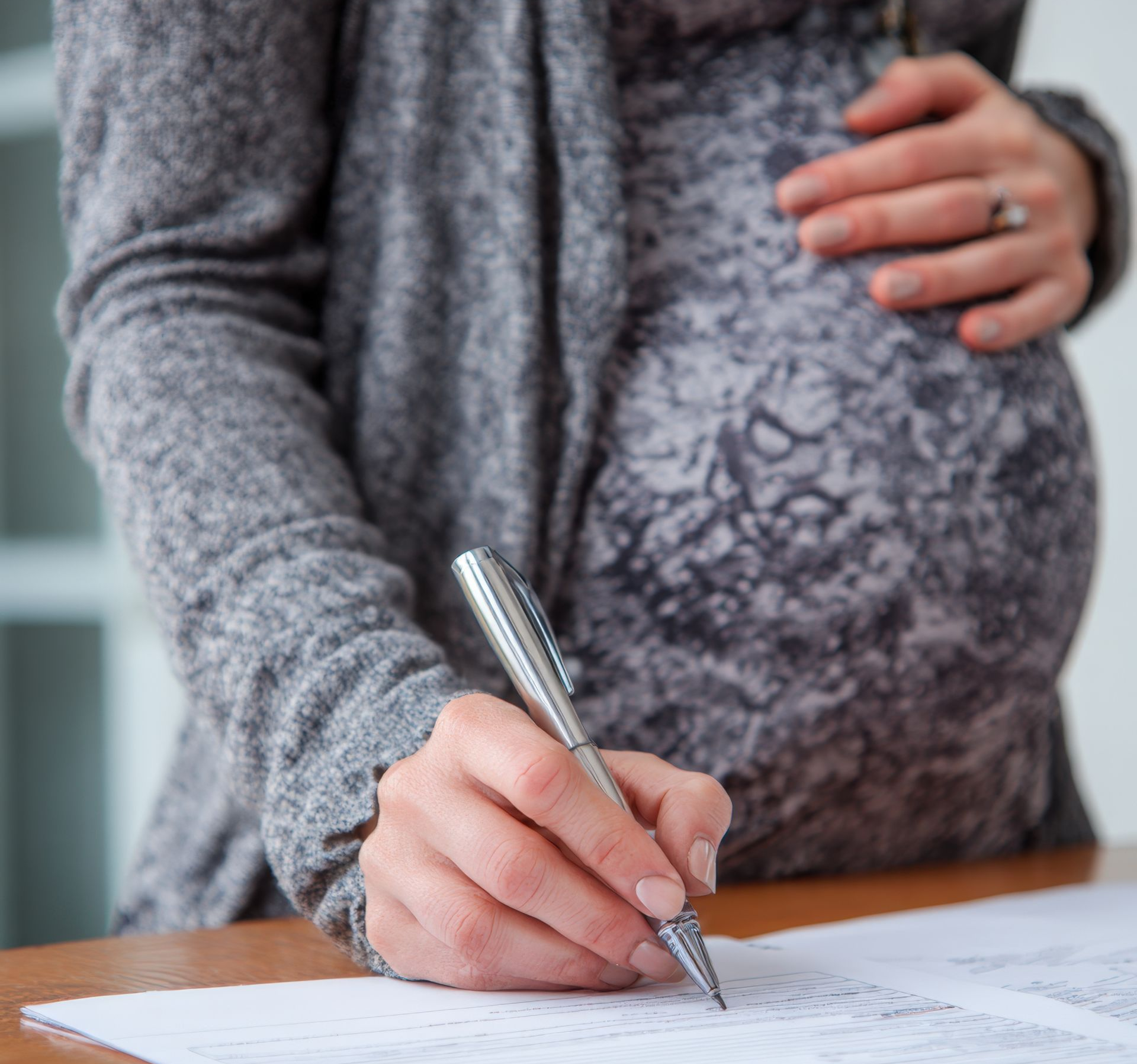 Une femme enceinte, vêtue d'un cardigan gris, écrit sur un document avec un stylo argenté, une main posée sur son ventre.