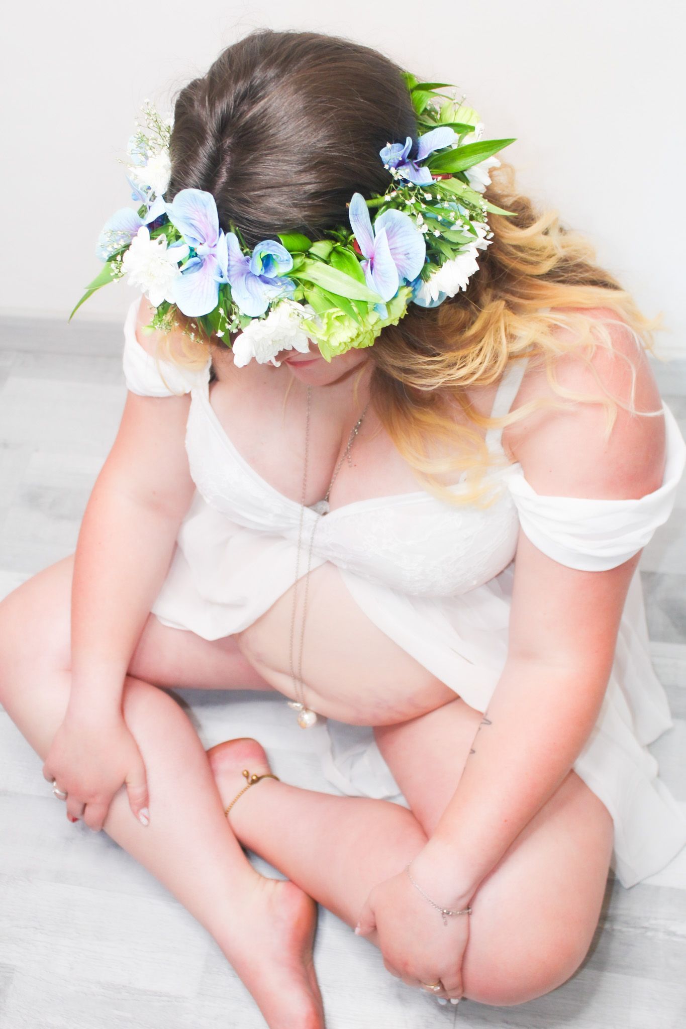 Une femme enceinte est assise par terre, vêtue d'une robe blanche et d'une couronne de fleurs bleues et blanches.