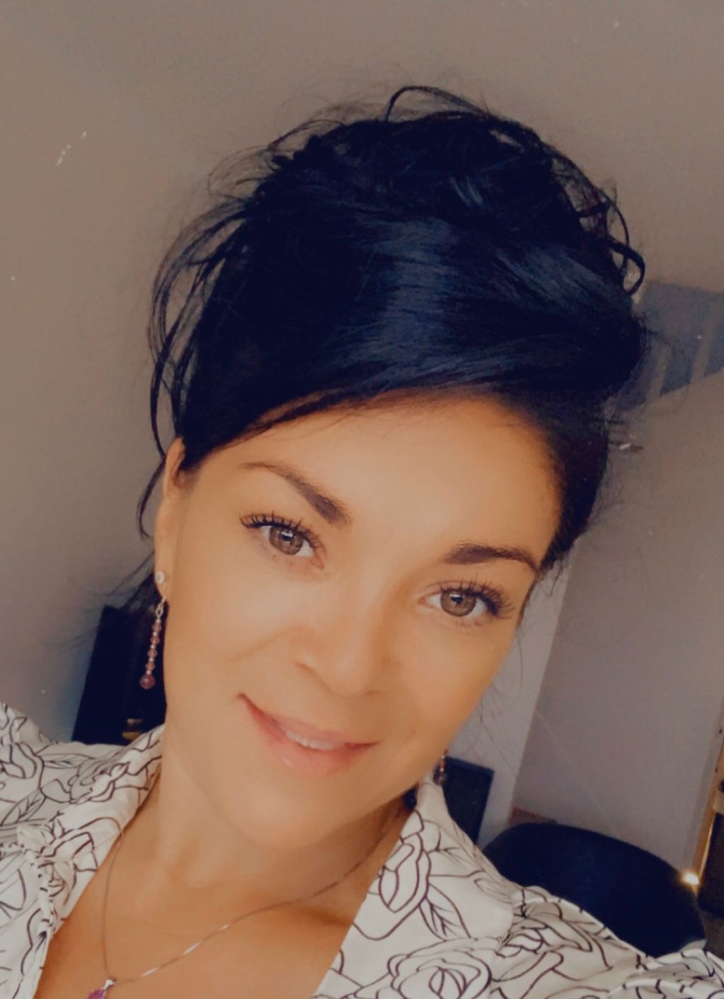 Une personne aux cheveux foncés coiffés en chignon regarde l'objectif, vêtue d'un haut blanc à motifs floraux noirs dessinés au trait.