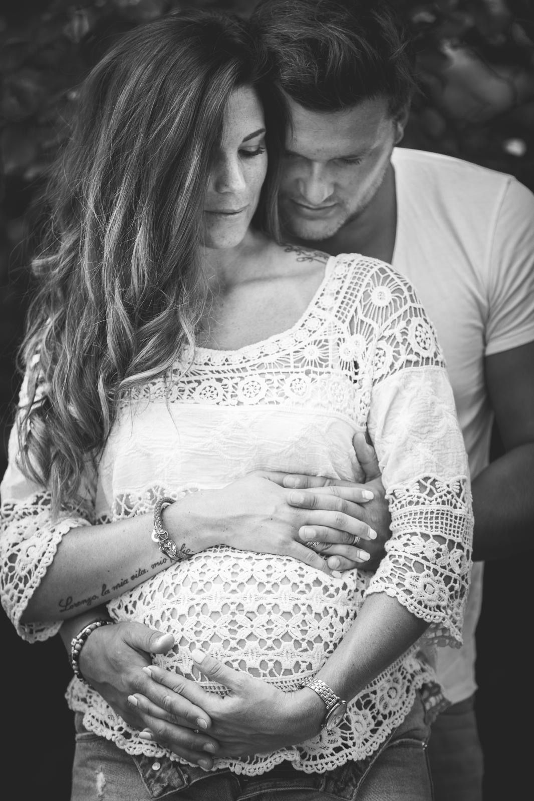 Une personne aux longs cheveux, vêtue d'un haut en dentelle, tient son ventre de femme enceinte tandis qu'une autre personne se tient derrière elle.