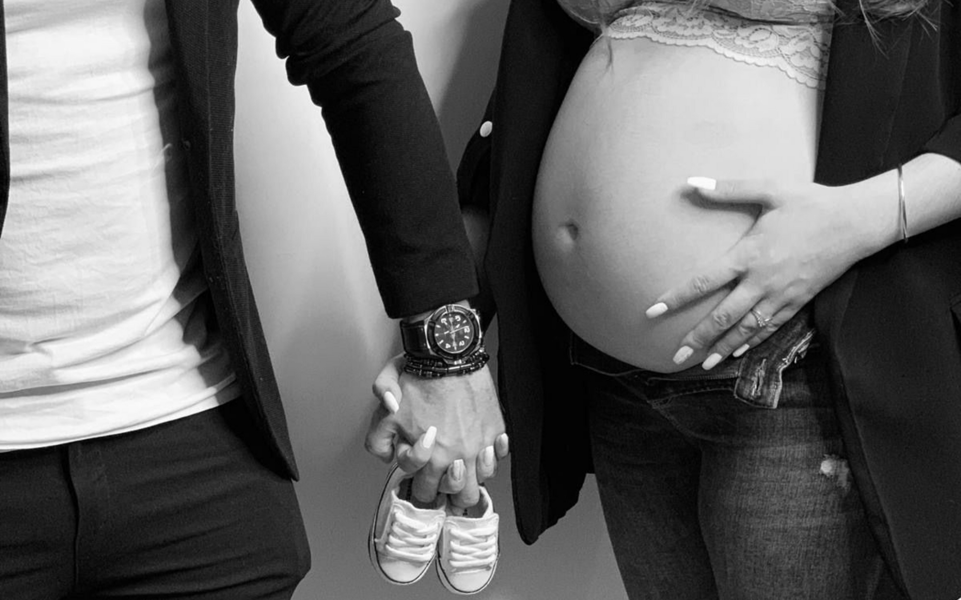 Une personne tenant de minuscules chaussures de bébé tout en touchant le ventre d'une femme enceinte, une autre personne lui tenant la main.