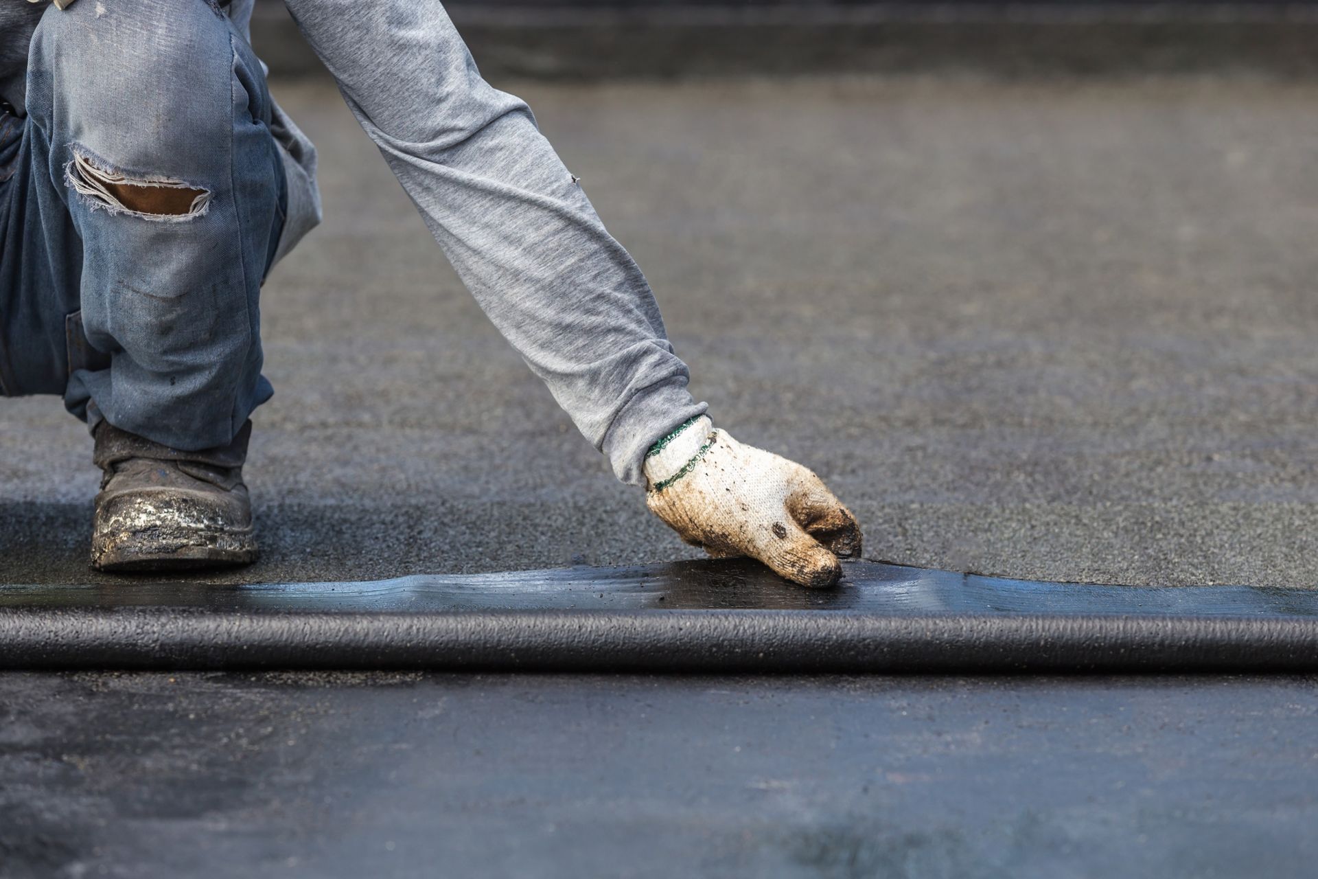 Una persona con guantes de trabajo se arrodilla sobre un techo plano, aplicando un material de techado negro.