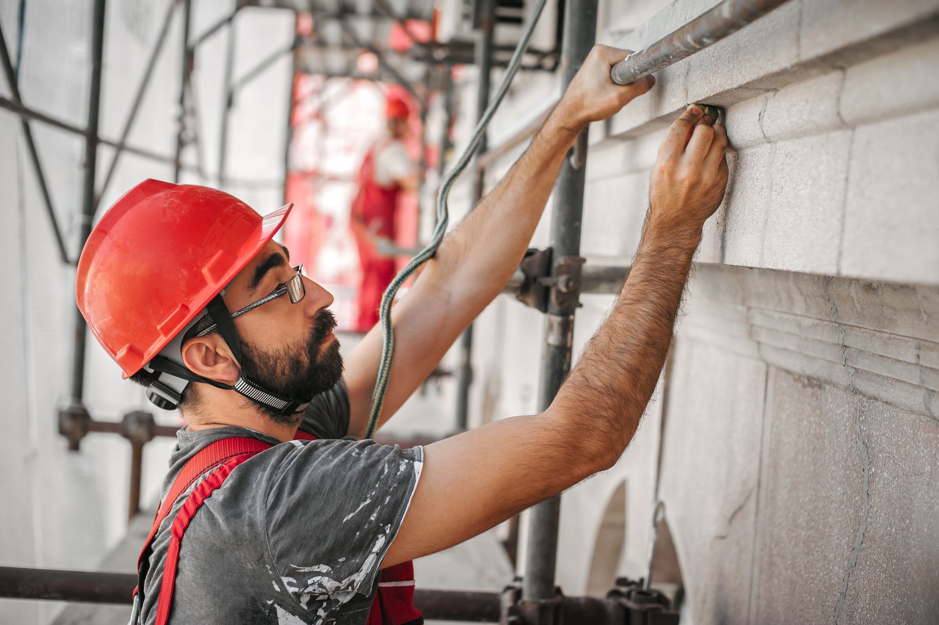 Trabajador de la construcción con casco rojo y arnés en un andamio, inspeccionando la fachada de un edificio.