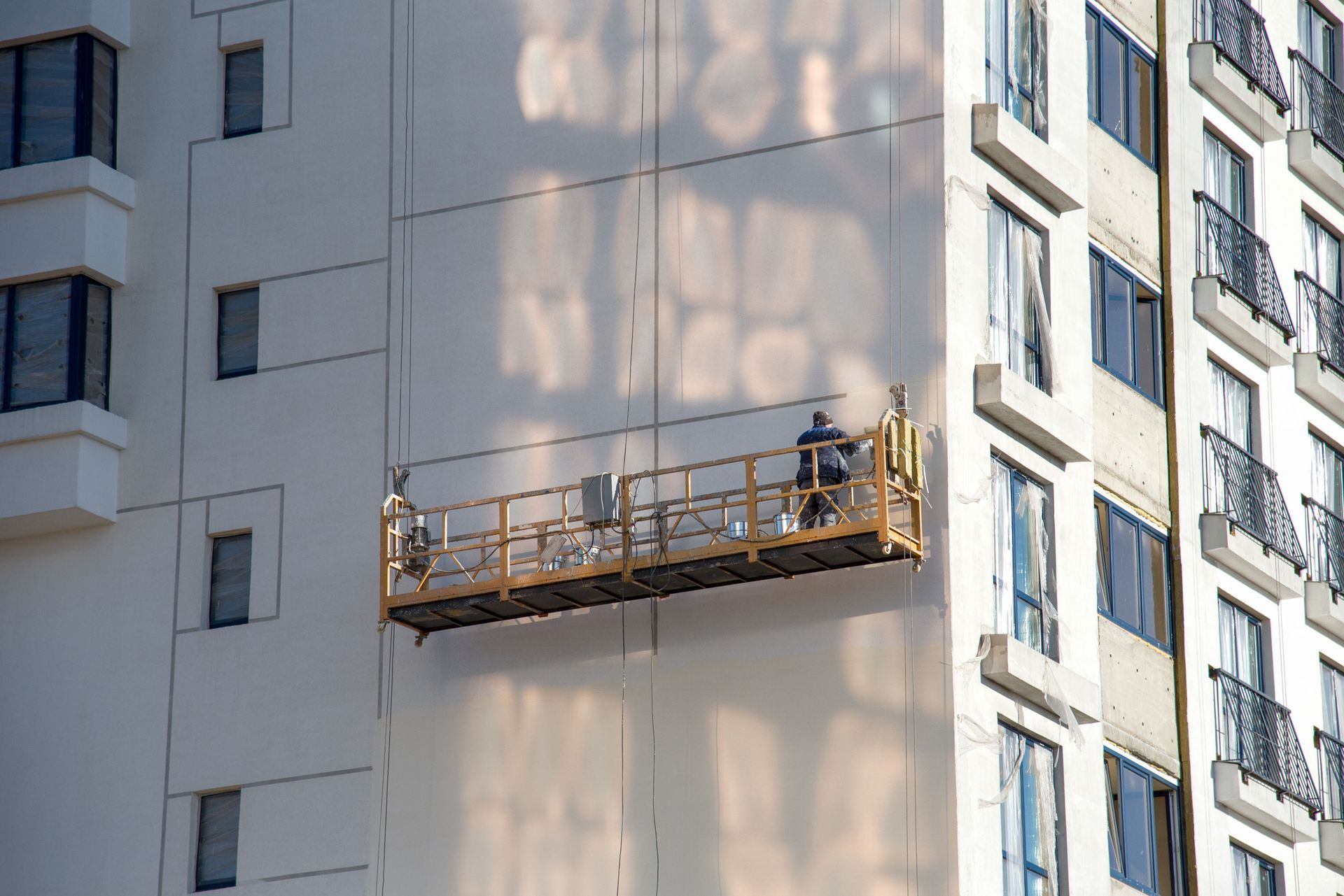 Obreros en una plataforma suspendida reparan la fachada de un edificio blanco. La luz del sol proyecta sombras en la pared.